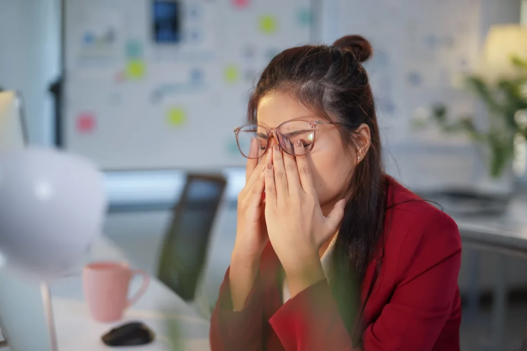 Woman with glasses rubbing her eyes at a computer due to blurred vision and eye strain
